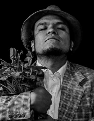 Retrato en blanco y negro del actor y director teatral Pedro Acevedo, con sombrero y saco a cuadros, sosteniendo un ramo de plantas entre sus brazos mientras mantiene los ojos cerrados en una pose introspectiva sobre un fondo negro.