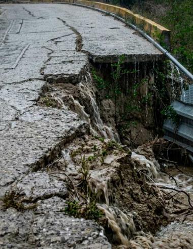 Fotografía de una carretera de concreto rural gravemente colapsada en un acantilado bajo una fuerte lluvia. Una parte significativa del pavimento se ha desmoronado. El agua de lluvia fluye activamente por las grietas y las caras expuestas de la tierra y la roca, y cae sobre un culvert de metal oxidado al pie del colapso. Árboles verdes se alinean en la colina bajo el clima lluvioso.