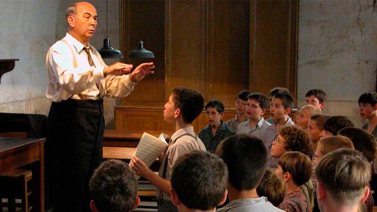 El profesor Clément Mathieu dirige un coro de niños en un salón de clases mientras varios alumnos cantan y observan atentamente durante un ensayo en una escena de la película francesa Los Coristas (Les Choristes), dirigida por Christophe Barratier.