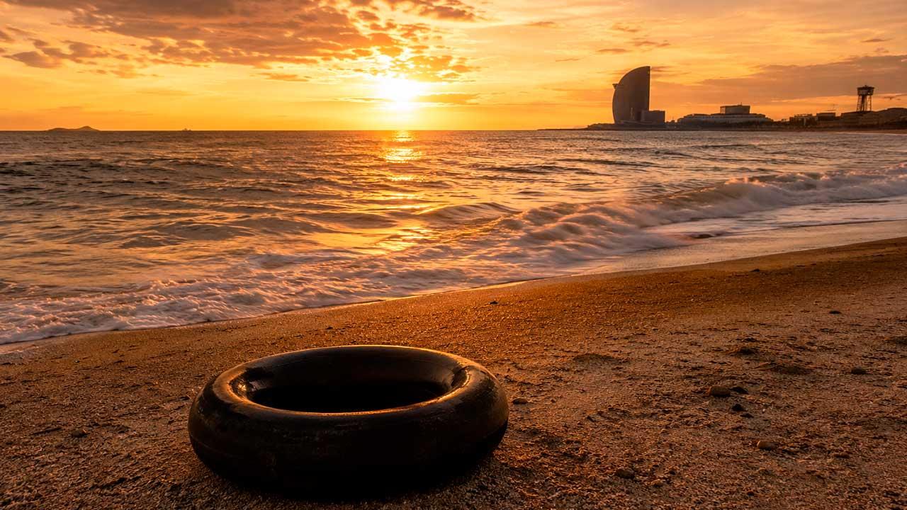 Una llanta de cámara de camión descansa sobre la arena de una playa al atardecer. El sol ilumina el horizonte con tonos dorados mientras el mar se extiende hacia una ciudad costera al fondo, evocando un paisaje de memoria, viaje y contemplación.