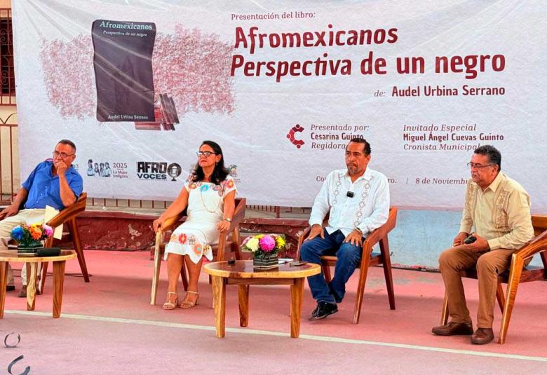 Audel Urbina Serrano presenta su libro "Afromexicanos, perspectiva de un negro" en Coyuca de Benítez, acompañado por la regidora Cesarina Quinto, el cronista Miguel Ángel Cuevas Quinto y otros invitados, frente al banner oficial del evento.
