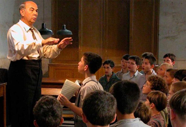 El profesor Clément Mathieu dirige un coro de niños en un salón de clases mientras varios alumnos cantan y observan atentamente durante un ensayo en una escena de la película francesa Los Coristas (Les Choristes), dirigida por Christophe Barratier.