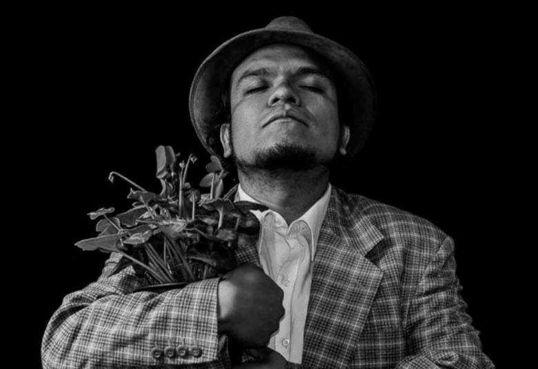 Retrato en blanco y negro del actor y director teatral Pedro Acevedo, con sombrero y saco a cuadros, sosteniendo un ramo de plantas entre sus brazos mientras mantiene los ojos cerrados en una pose introspectiva sobre un fondo negro.