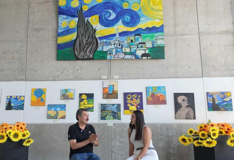Entrevista a la Maestra Iliana Fuentes en la Sala de Exposiciones del Fuerte de San Diego, rodeada de pinturas de girasoles y réplicas de obras de Vincent van Gogh hechas por estudiantes.