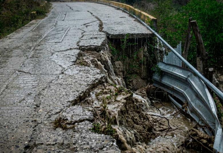 Fotografía de una carretera de concreto rural gravemente colapsada en un acantilado bajo una fuerte lluvia. Una parte significativa del pavimento se ha desmoronado. El agua de lluvia fluye activamente por las grietas y las caras expuestas de la tierra y la roca, y cae sobre un culvert de metal oxidado al pie del colapso. Árboles verdes se alinean en la colina bajo el clima lluvioso.
