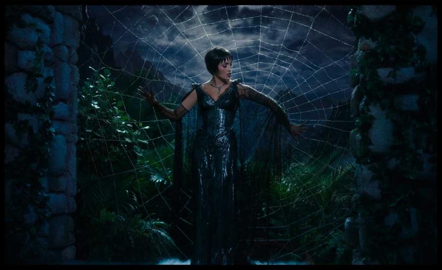 Jennifer López como La Mujer Araña en un escenario teatral, de pie frente a una telaraña gigante que refleja su silueta, vestida con un traje brillante de lentejuelas.
