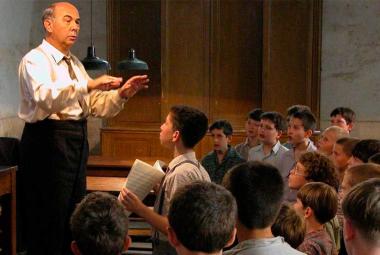 El profesor Clément Mathieu dirige un coro de niños en un salón de clases mientras varios alumnos cantan y observan atentamente durante un ensayo en una escena de la película francesa Los Coristas (Les Choristes), dirigida por Christophe Barratier.
