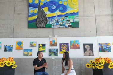 Entrevista a la Maestra Iliana Fuentes en la Sala de Exposiciones del Fuerte de San Diego, rodeada de pinturas de girasoles y réplicas de obras de Vincent van Gogh hechas por estudiantes.
